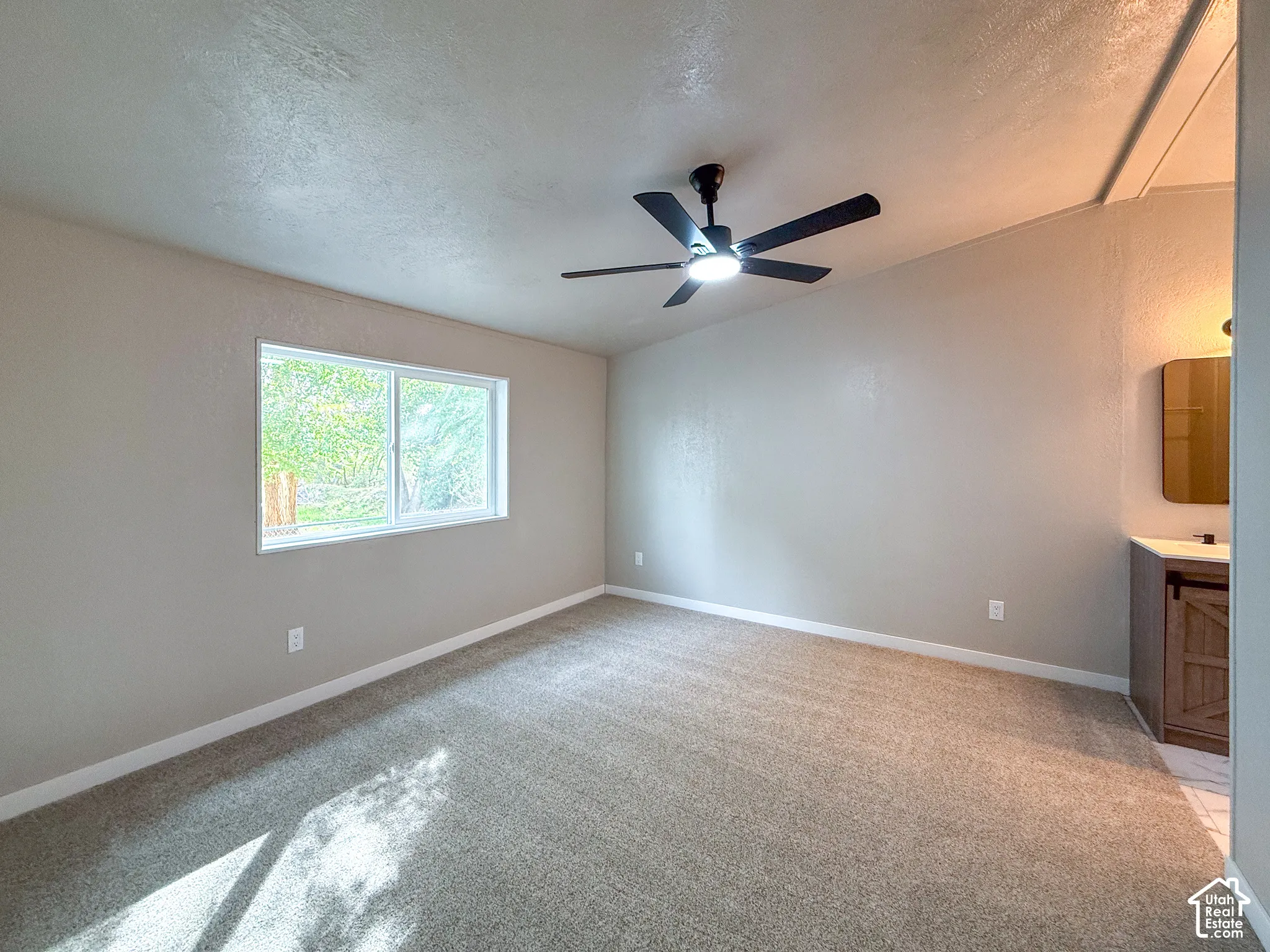 Neutral-toned wall finishes, overhead ceiling fan with integrated lighting, window with exterior landscaping, carpeted flooring, and a built-in vanity with a rectangular mirror