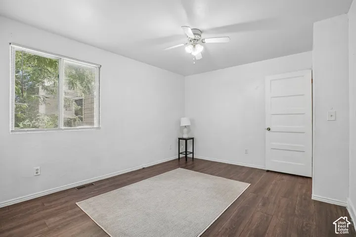 Wood-finish flooring, white walls, five-panel interior door, window with blinds, and ceiling fan with integrated lighting