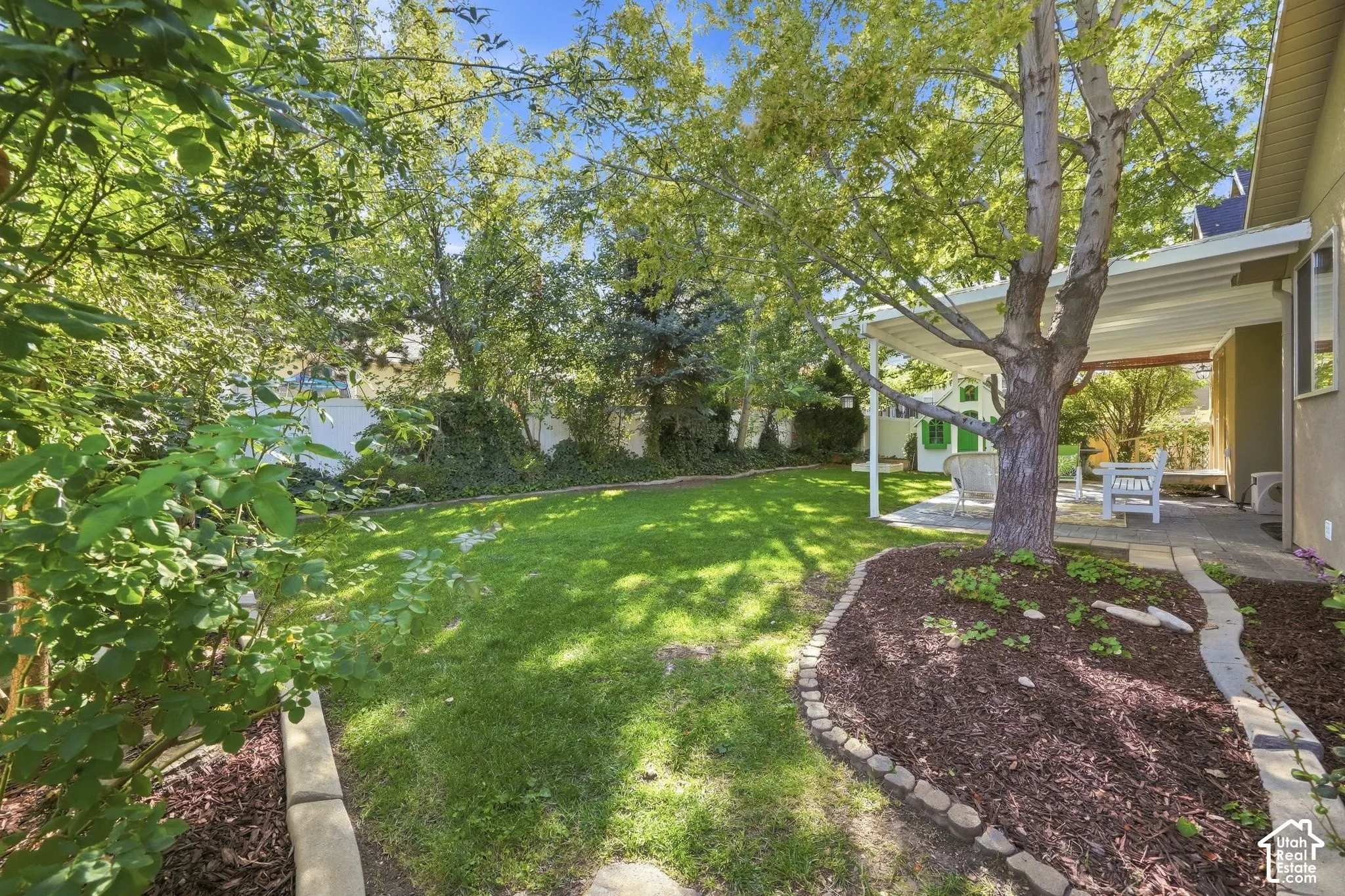 Expansive green lawn, mature trees, covered patio, mulched garden beds with stone edging, and paved patio area