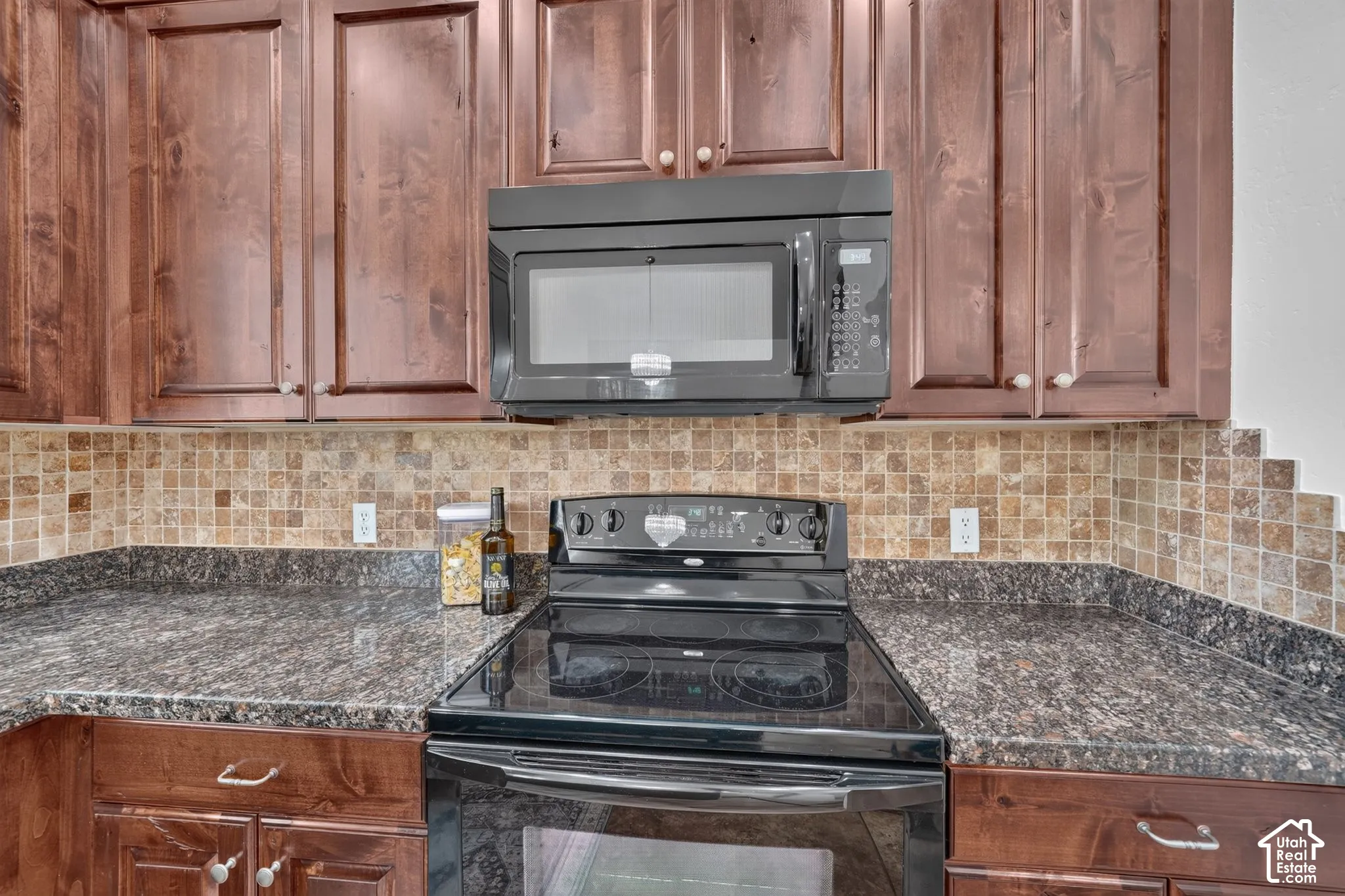 Dark wood-finish cabinetry, black over-the-range microwave, tile backsplash, and dark speckled countertops