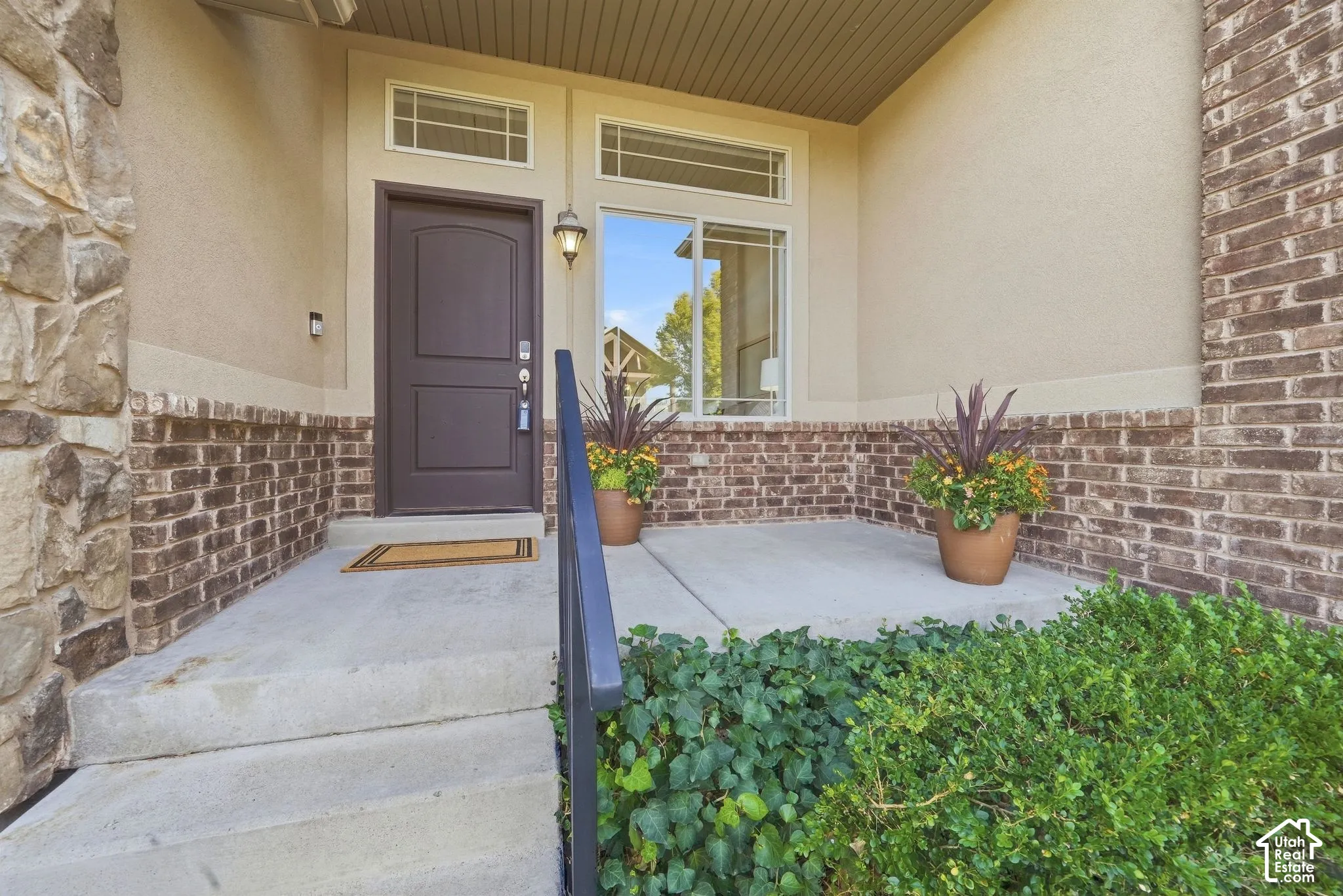 Entryway featuring a dark paneled door with transom windows, brick and stone facade, concrete porch with steps, and a metal handrail
