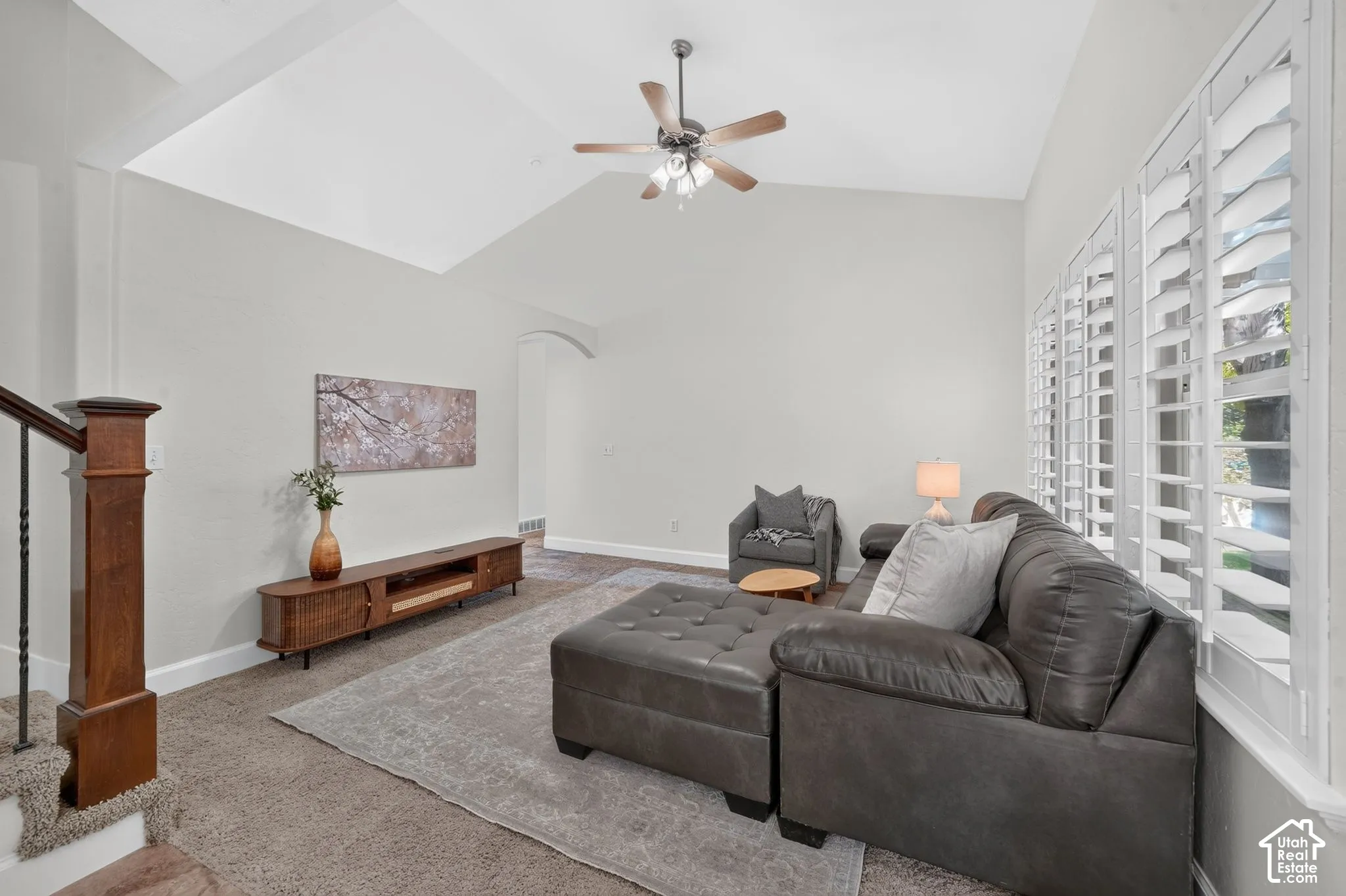 Vaulted ceiling with a ceiling fan, white plantation shutters, dark wood-finish newel post with wrought iron spindles, and a light gray wall finish