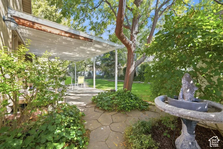 Paved stone patio featuring a white pergola structure, mature trees, manicured lawn, and a stone birdbath