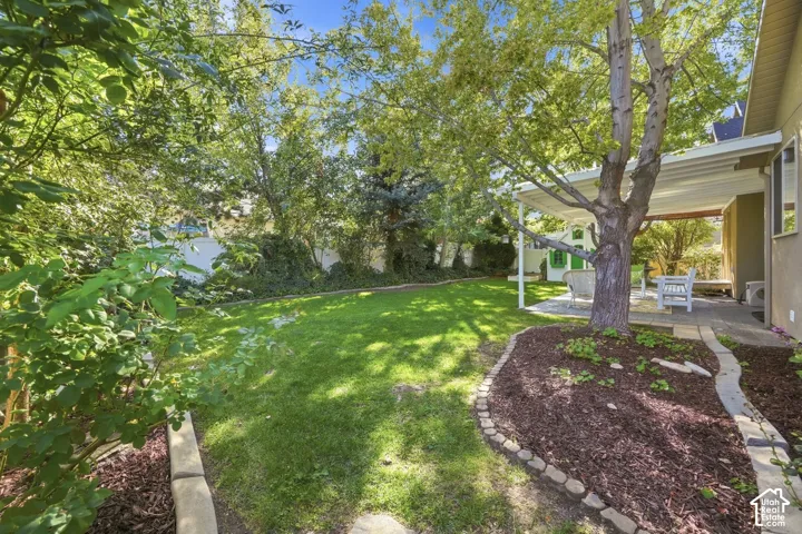 Expansive green lawn, mature trees, covered patio, mulched garden beds with stone edging, and paved patio area