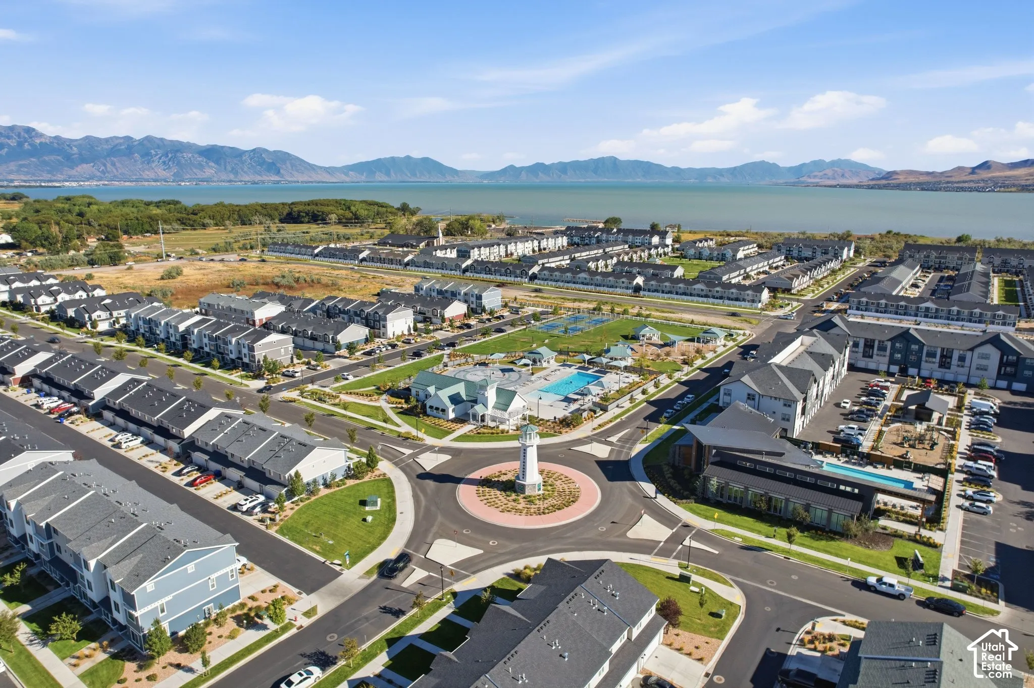 Community featuring multiple residential structures, a central roundabout with a lighthouse-style structure, a pool area, sports courts, and a large body of water in the background