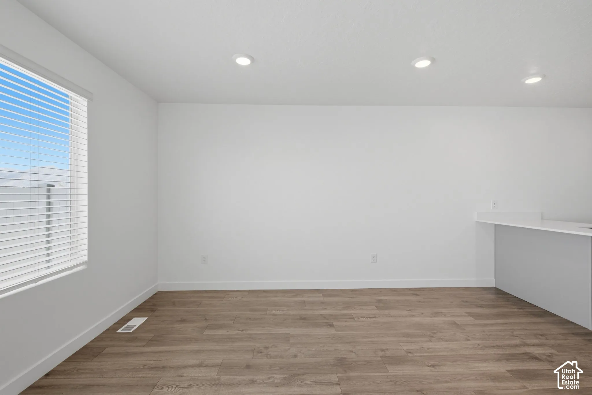 Wood-finish flooring, recessed ceiling lighting, a large window with blinds, and a built-in counter surface with an overhang