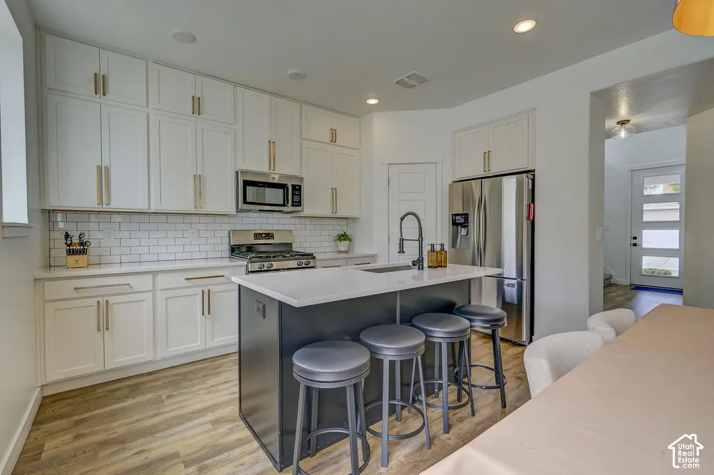 Kitchen featuring white shaker-style cabinetry, subway tile backsplash, stainless steel appliances, a center island with countertop seating, and wood-finish flooring