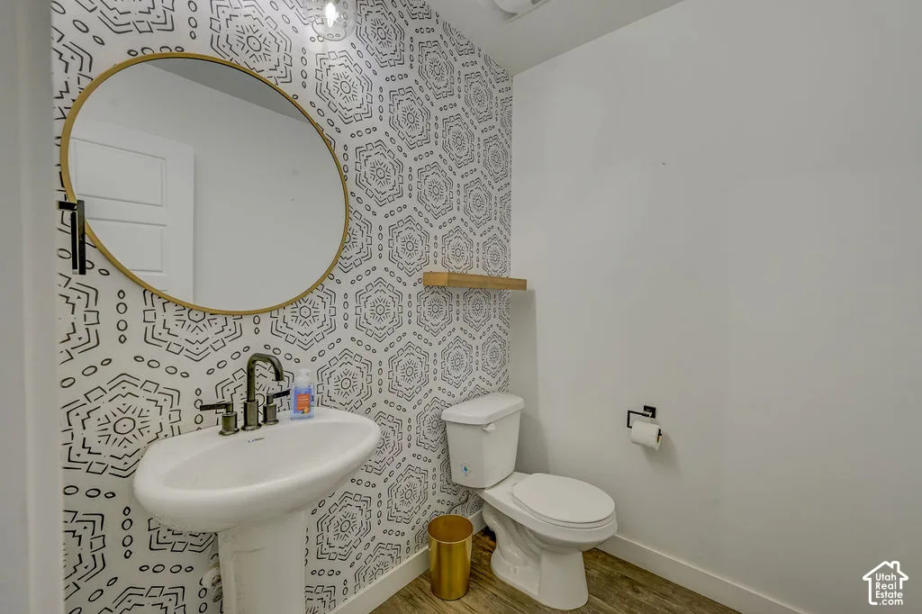 Pedestal sink with dark-toned fixtures, round framed mirror, patterned accent wall, white toilet, and wood-finish flooring