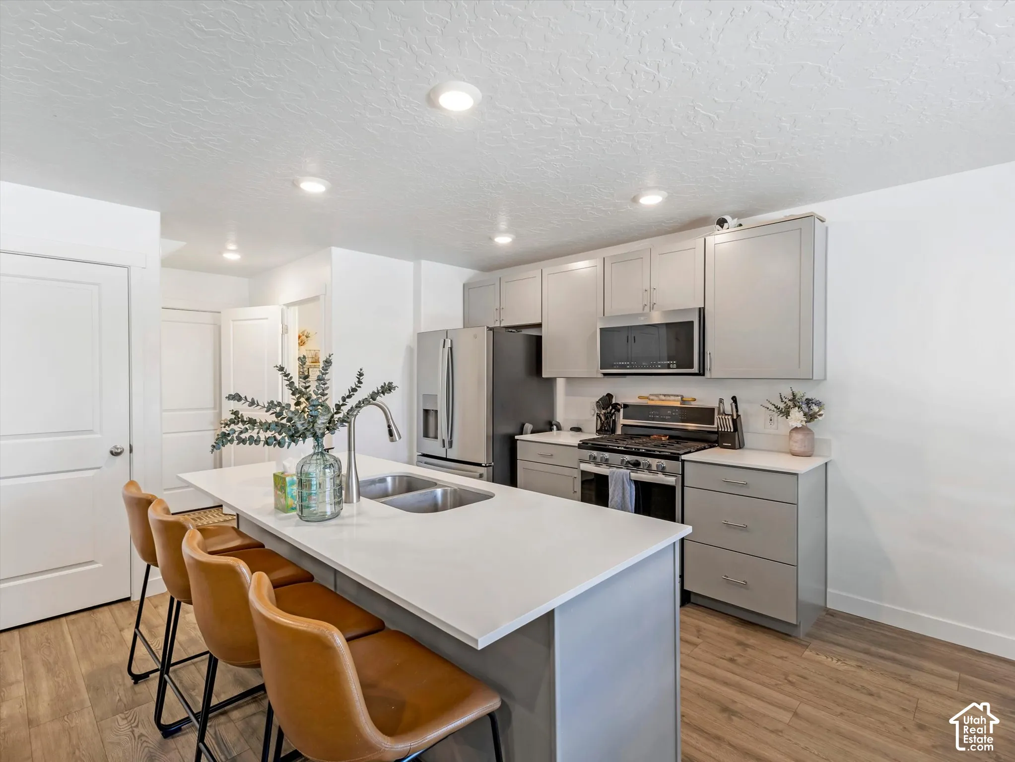 Kitchen featuring a large central island with an integrated sink, light grey cabinetry, stainless steel appliances, and wood-finish flooring