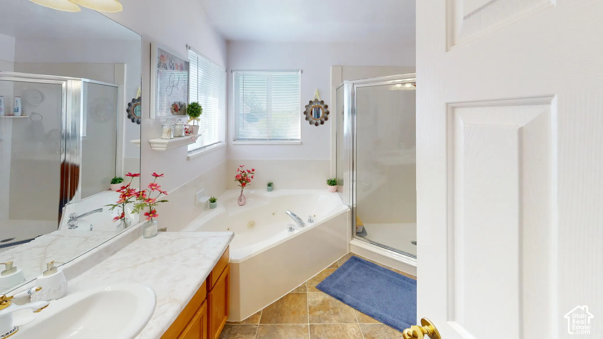 Bathroom featuring a corner jetted tub, glass-enclosed shower, dual windows with blinds, and a vanity with a large mirror
