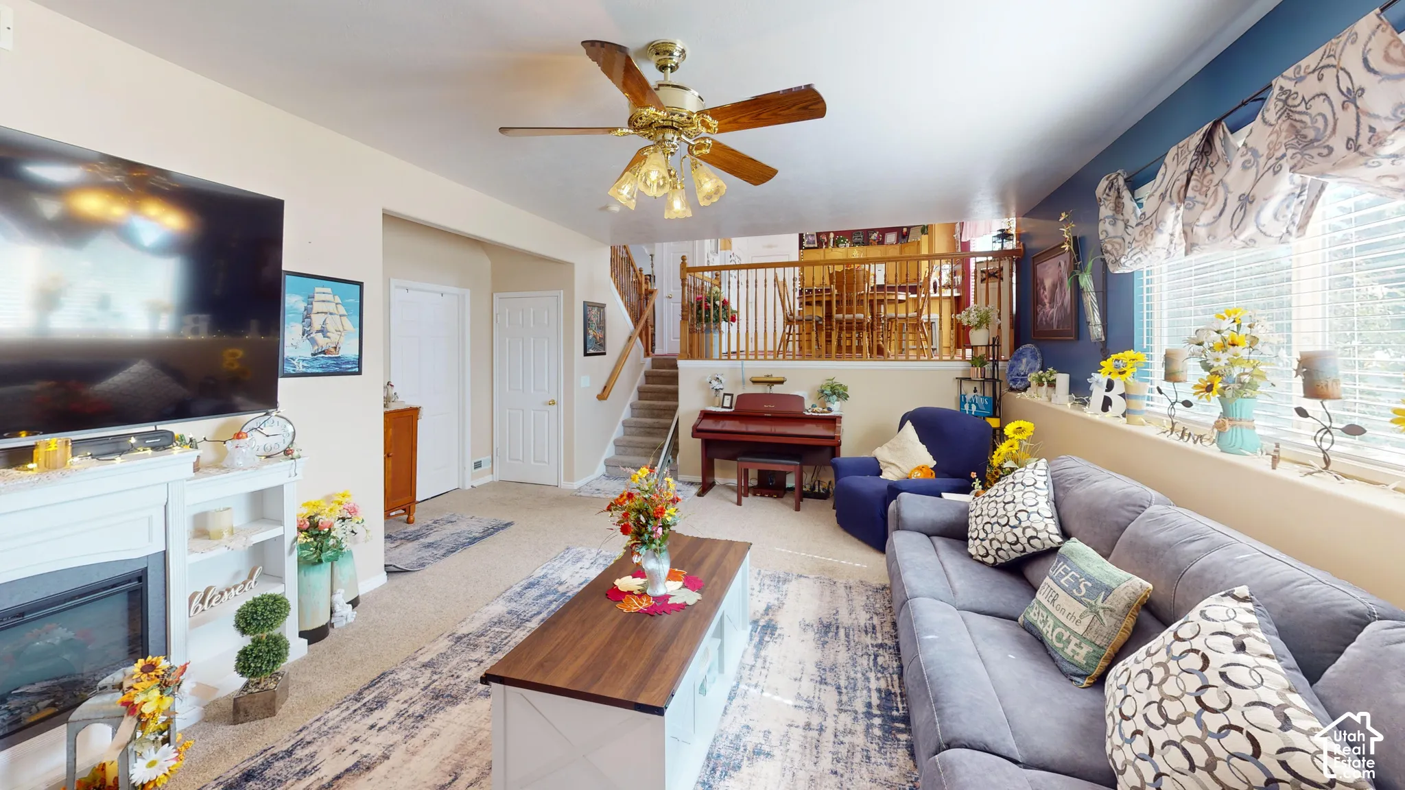 Recessed fireplace with white mantel and shelving, ceiling fan with integrated lighting, wood-finish staircase with white risers, window with blinds, and white interior doors