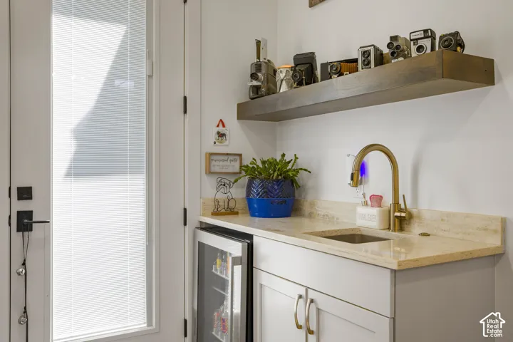 White cabinetry with brushed nickel hardware, stone countertop with integrated sink, brass-finish gooseneck faucet, built-in beverage refrigerator, and floating wood shelf