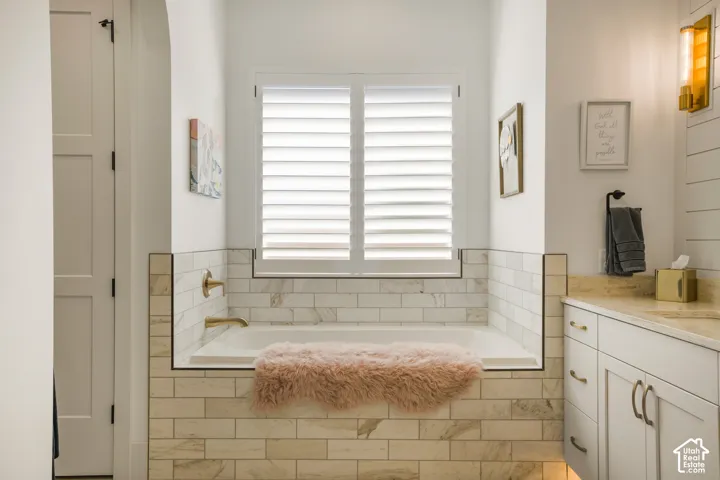 Built-in soaking tub with subway tile surround, plantation shutters, and a vanity with a stone countertop