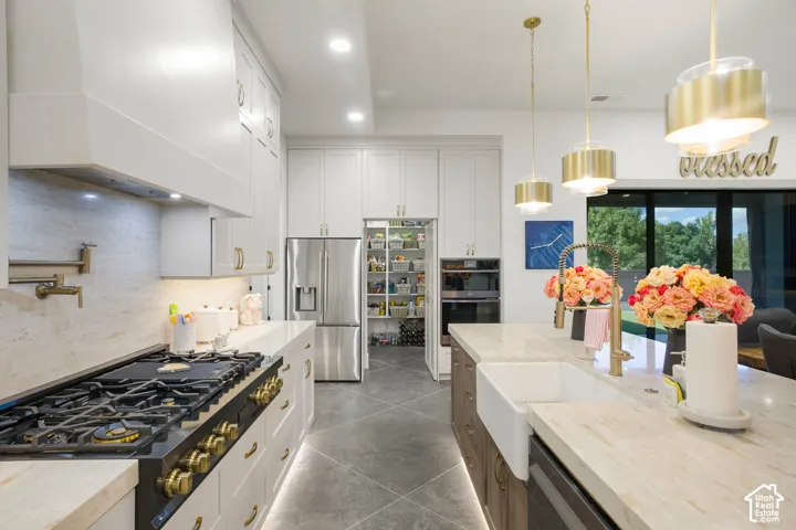 White cabinetry with gold-tone hardware, professional-grade range, stainless steel appliances, large kitchen island with farmhouse sink, and contemporary pendant lighting