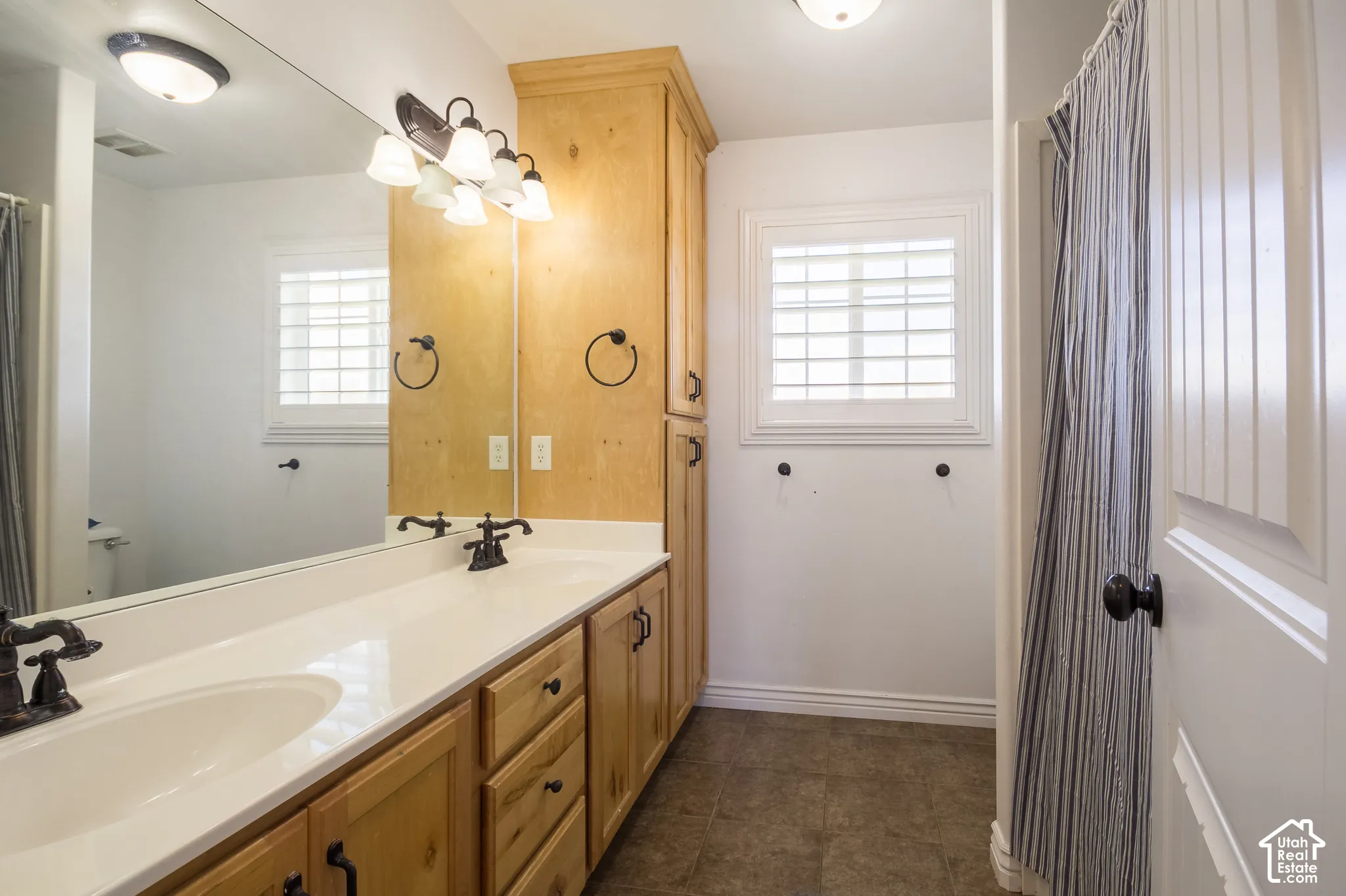 Double vanity with integrated sinks and dark bronze faucets, wood cabinetry, large mirror, and window with plantation shutters