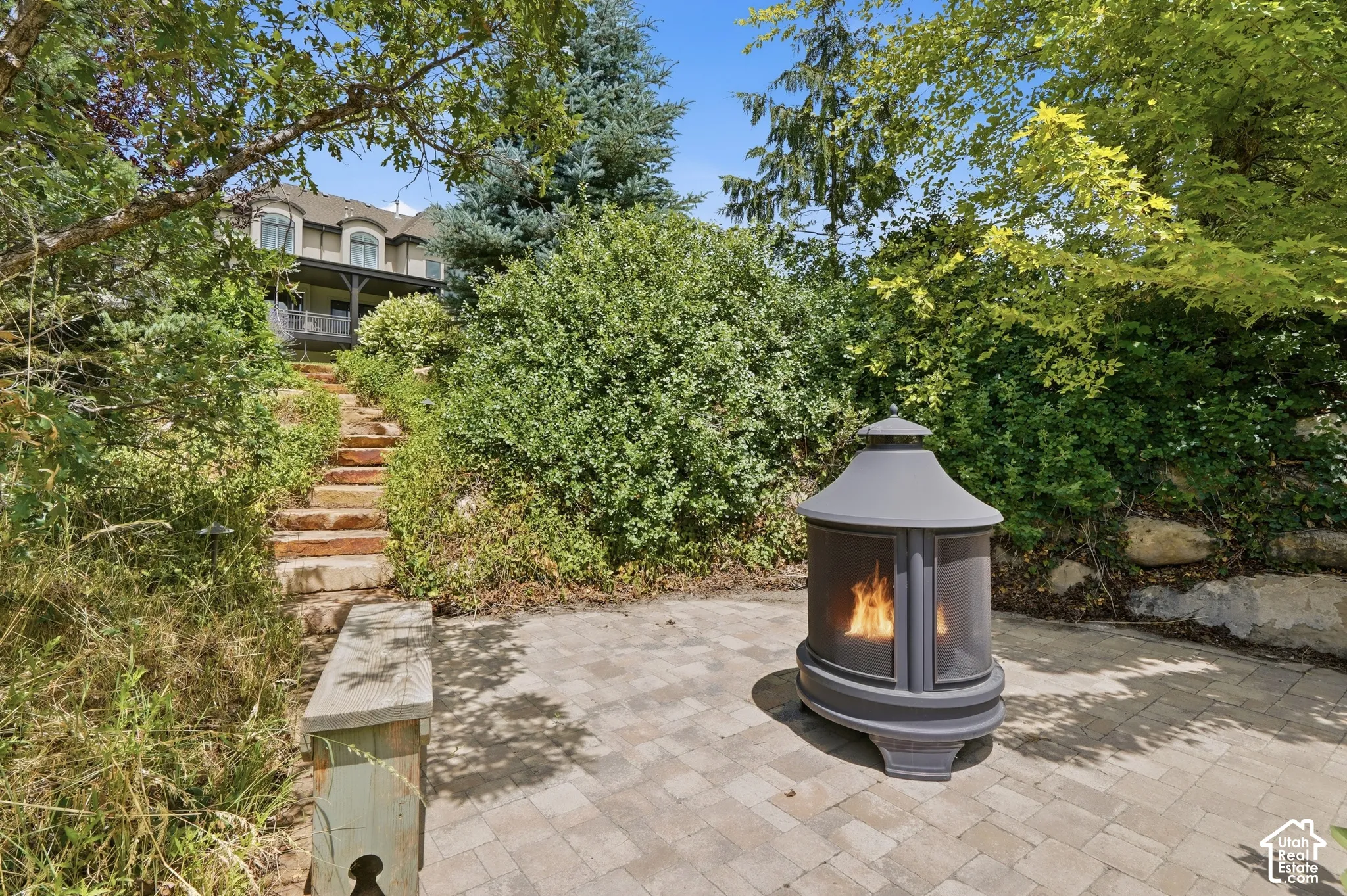 Paved patio area, stone retaining wall, stone steps, lush landscaping, and a multi-level structure in the background
