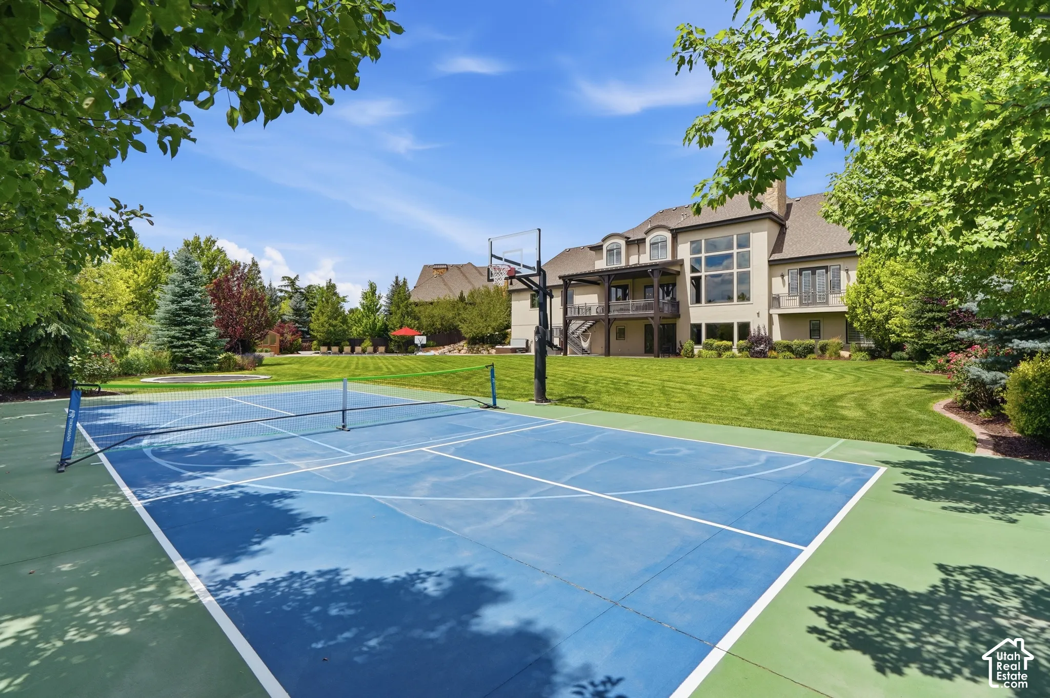 Dedicated sport court with basketball hoop and tennis net, expansive green lawn, mature trees, multi-story exterior with large windows, and a covered balcony