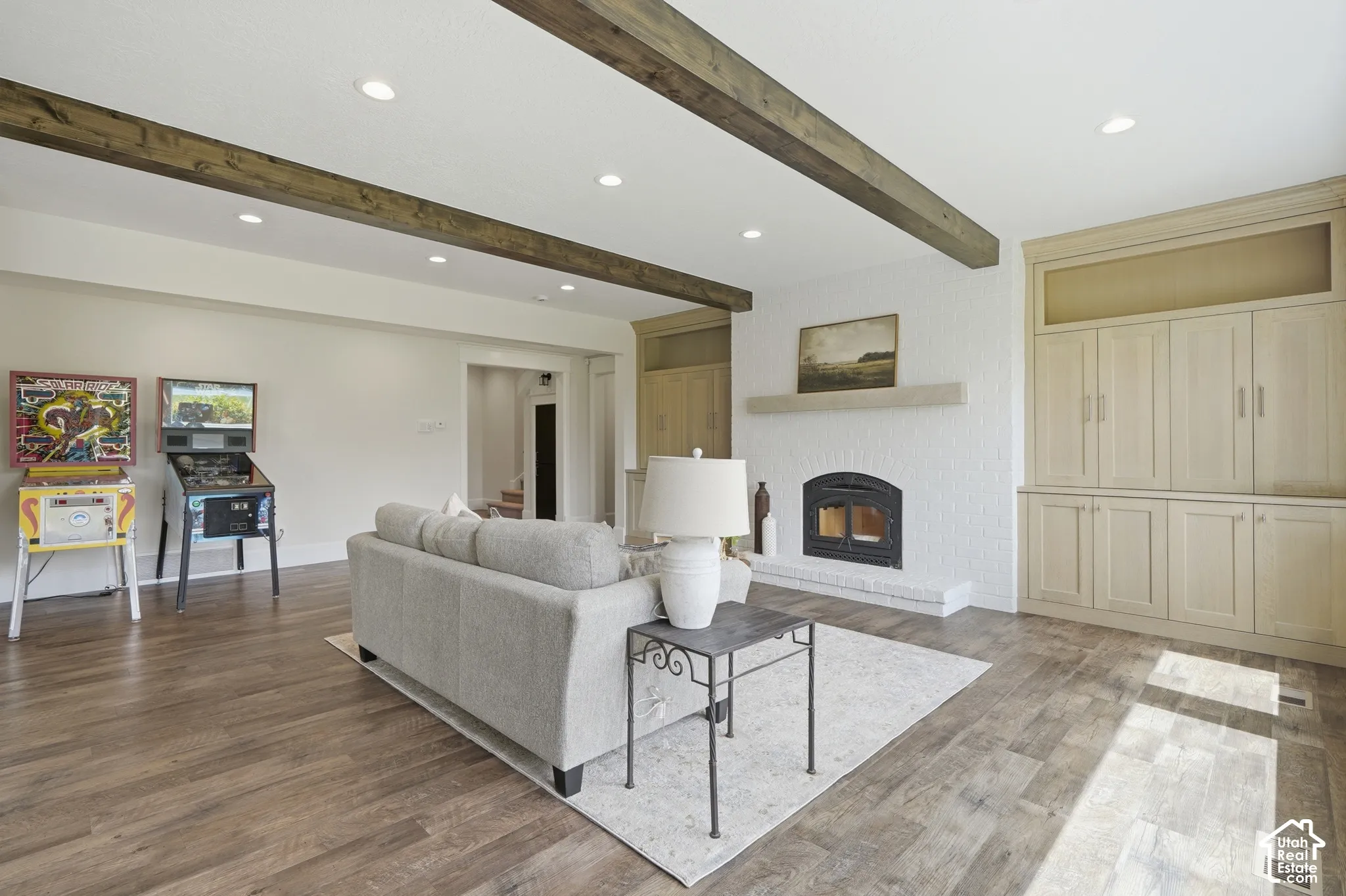 Exposed wood ceiling beams, recessed lighting, wood-finish flooring, a white brick fireplace with a stone mantel, and built-in cabinetry