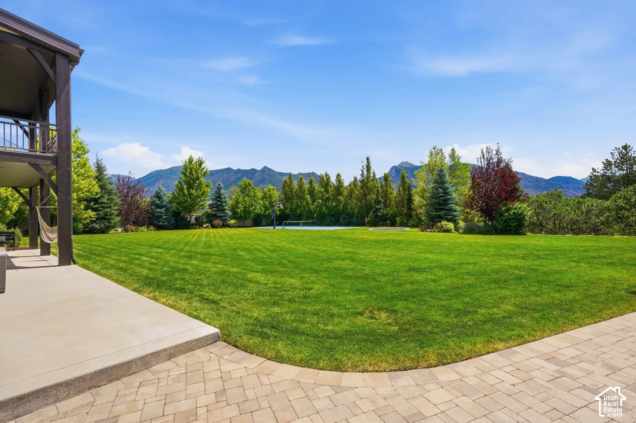 Expansive lawn with mature trees and a sports court, framed by mountain scenery, featuring a lower-level patio and an upper-level balcony