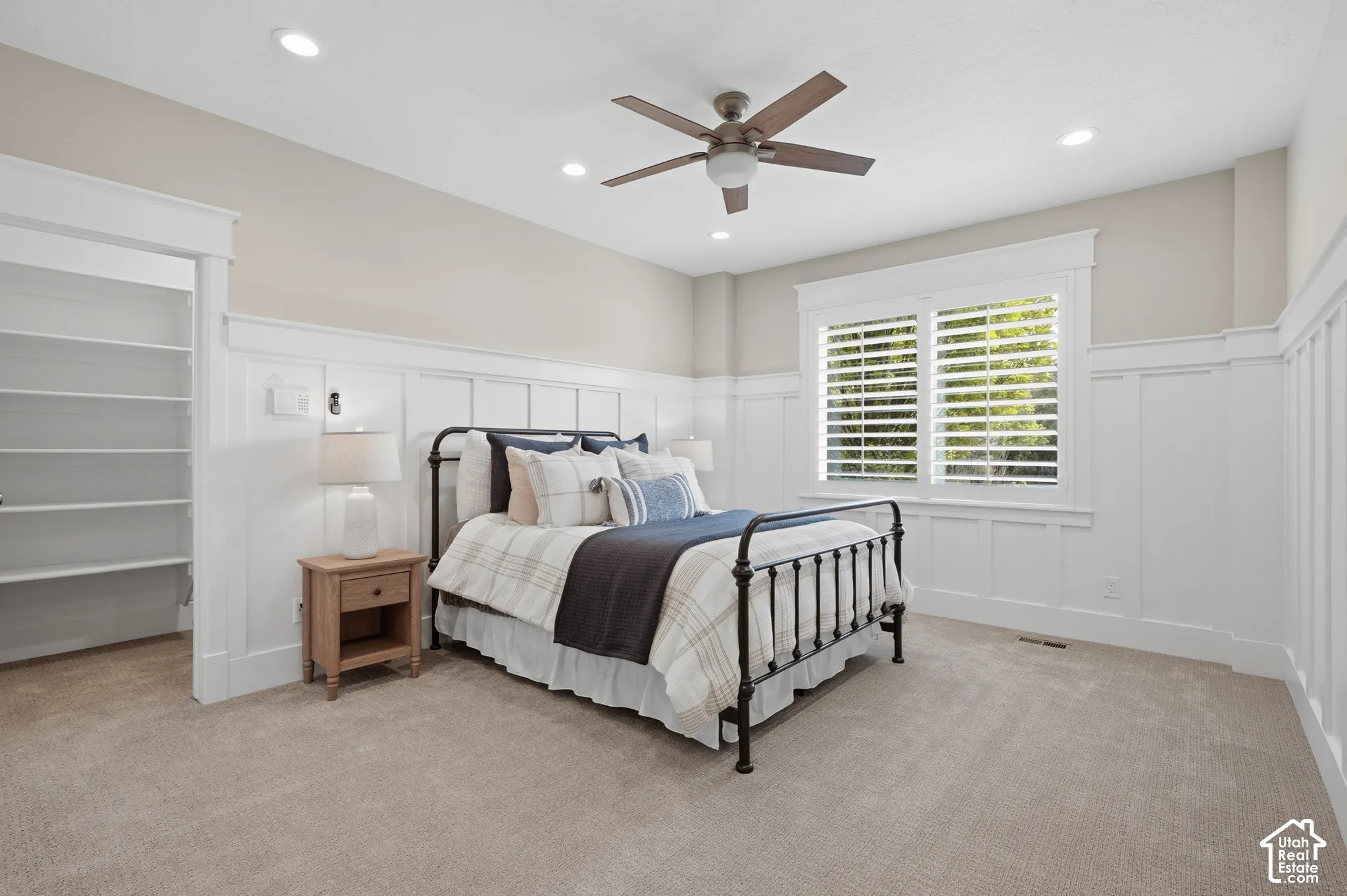 Recessed ceiling lighting, ceiling fan, white wainscoting, built-in shelving, and window with plantation shutters