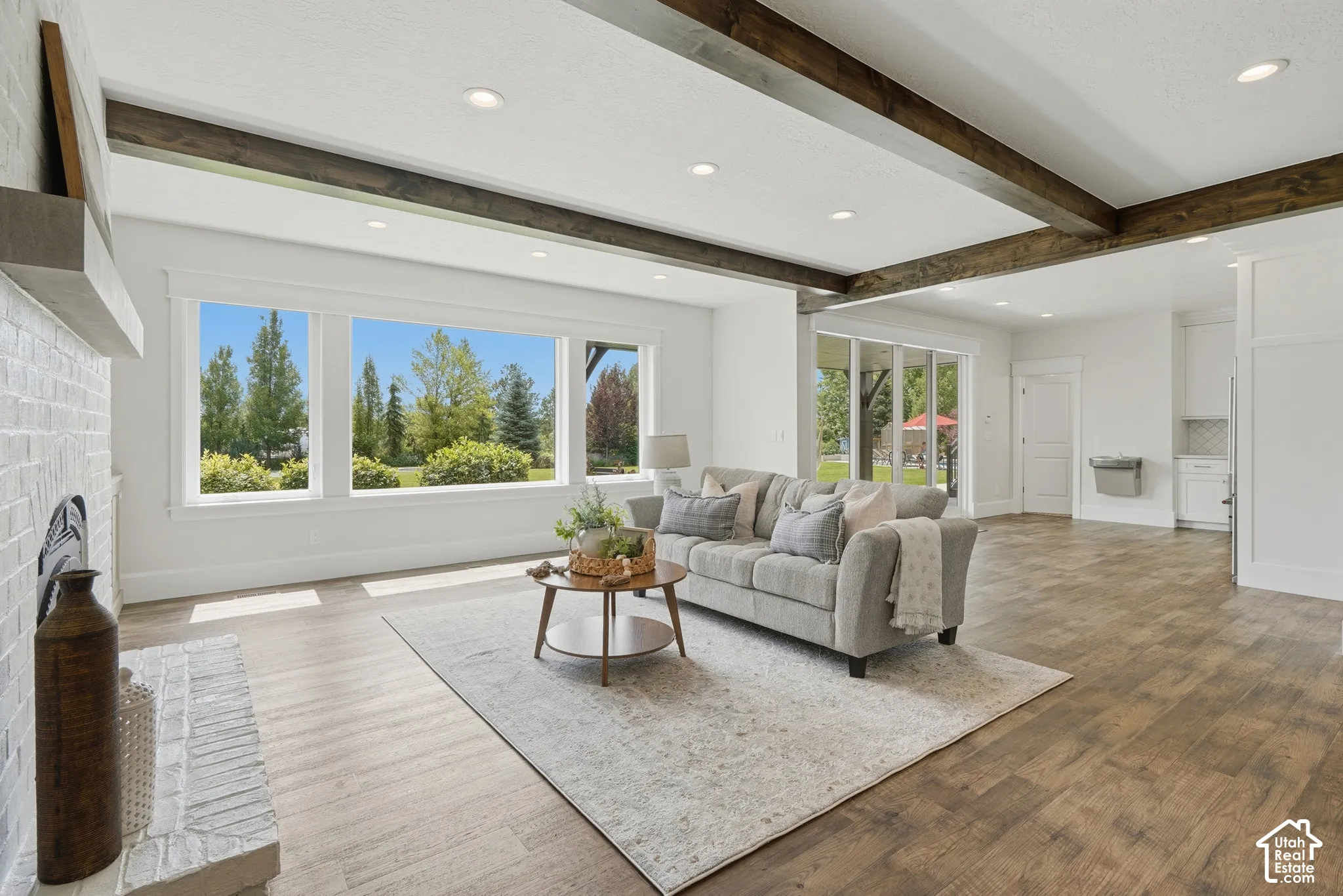 Expansive living space featuring wood-finish flooring, exposed ceiling beams, a white brick fireplace, recessed lighting, and large windows