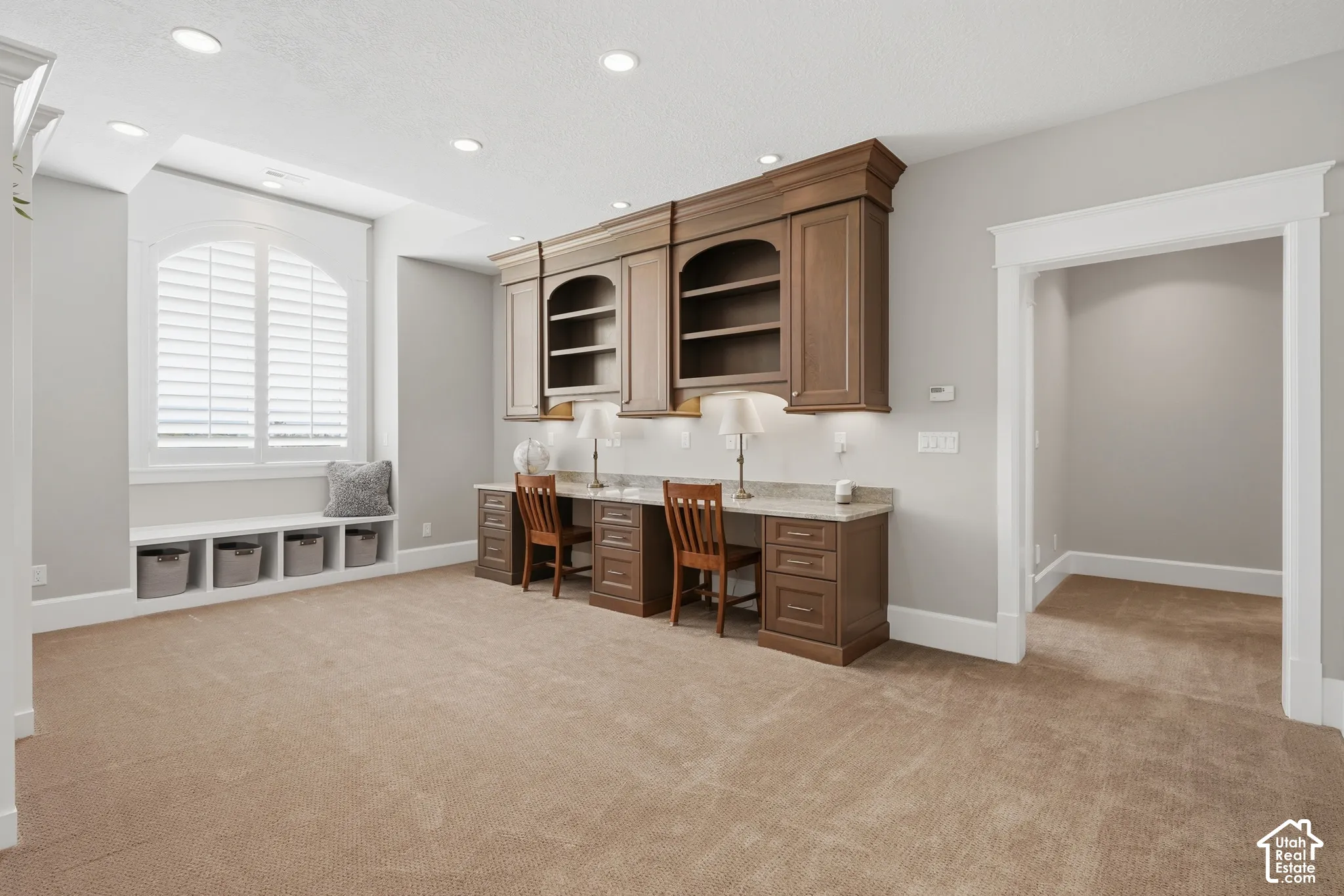 Built-in desk with overhead cabinetry, recessed lighting, carpet flooring, arched window with plantation shutters, and an integrated window bench with open storage
