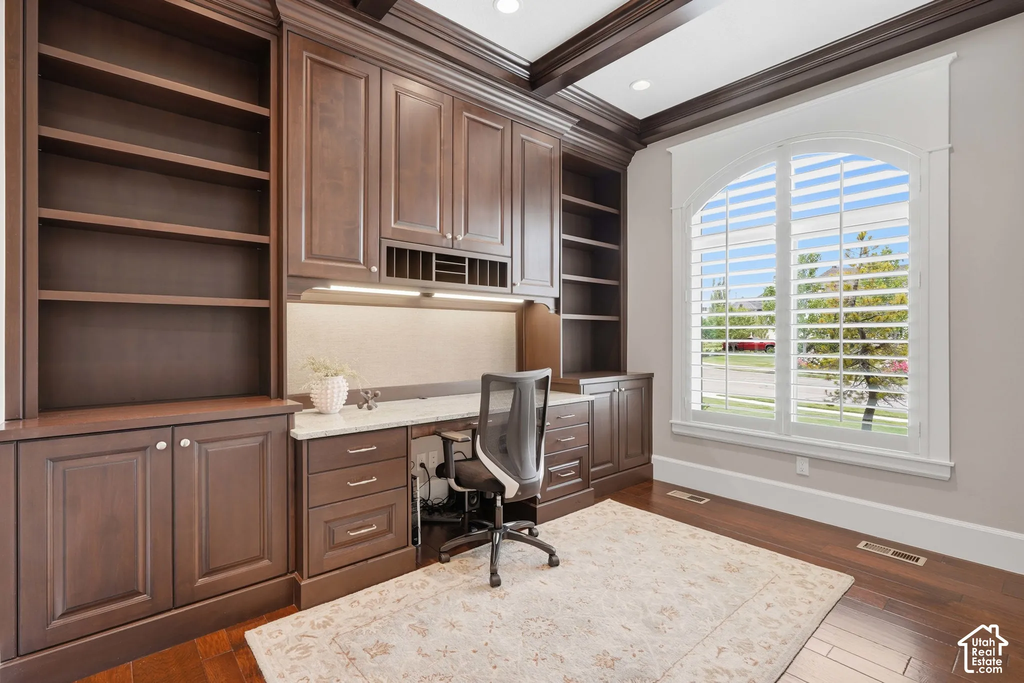 Custom built-in cabinetry and shelving, coffered ceiling, wood-finish flooring, arched window with plantation shutters, integrated desk lighting