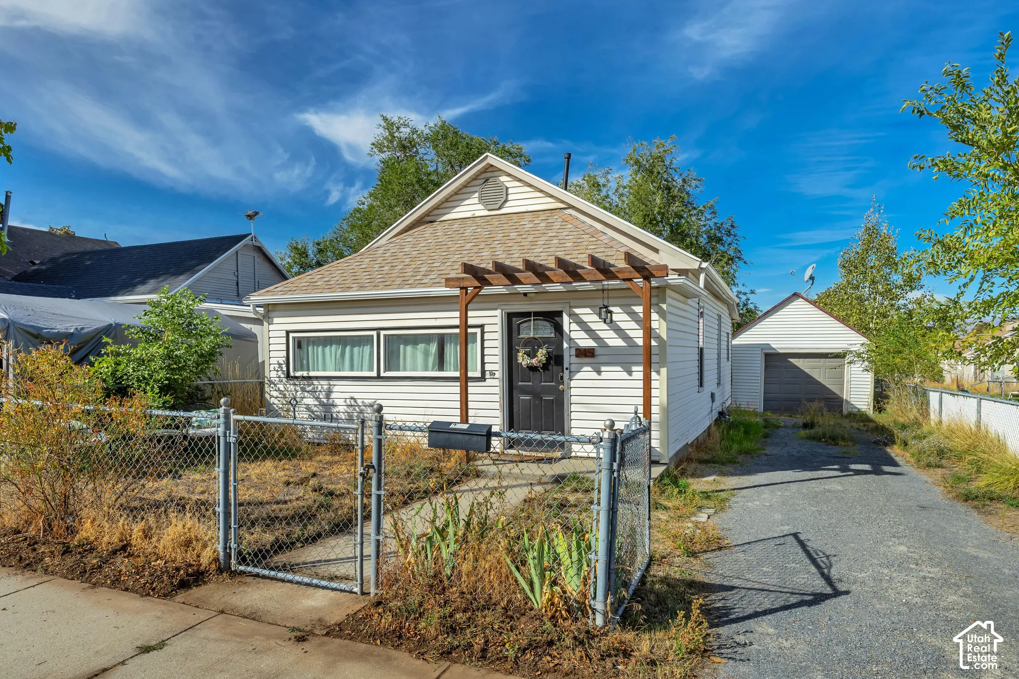 White siding exterior with a pitched roof, front entry with an overhead pergola, chain-link front yard fencing, and an asphalt driveway leading to a detached garage