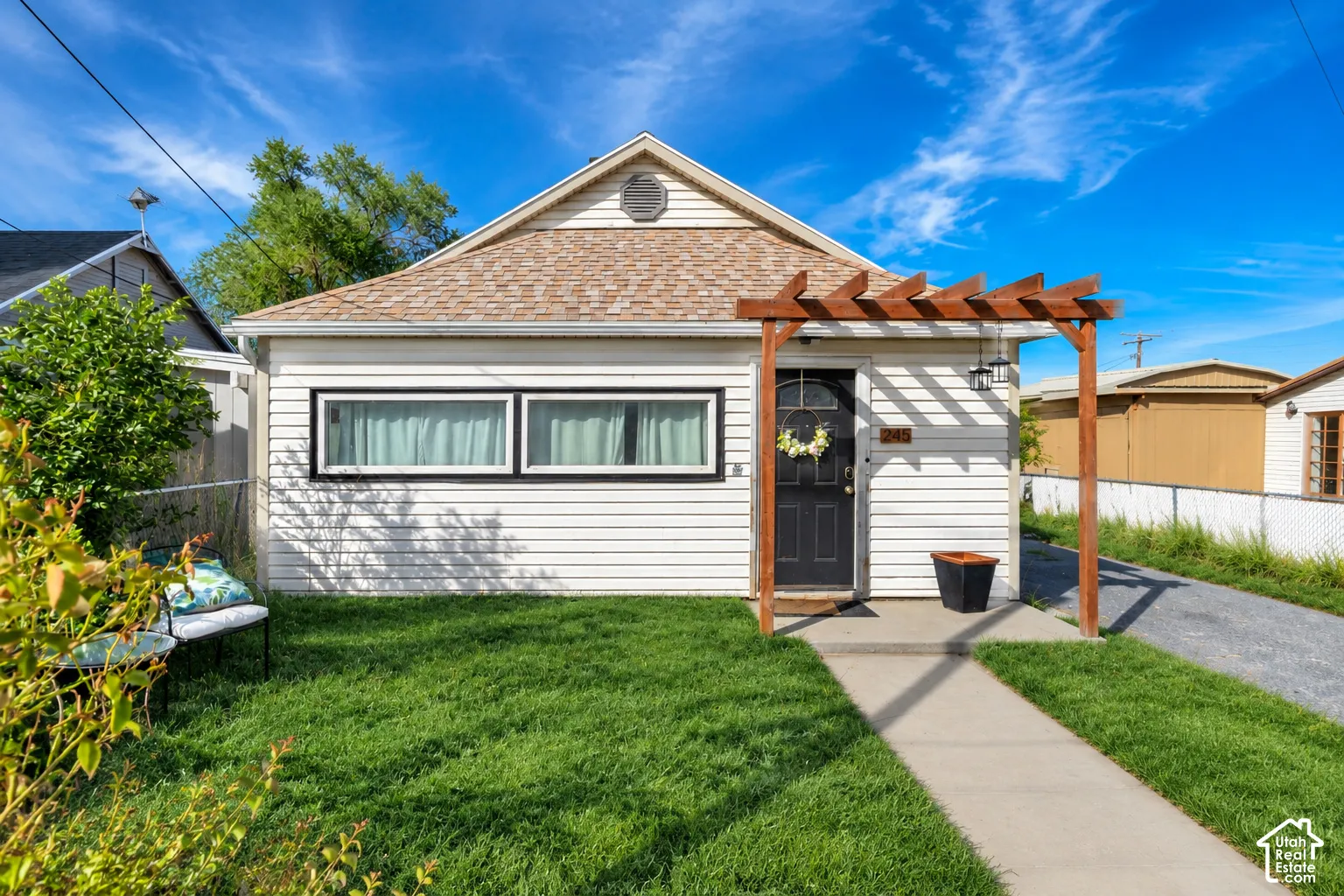 White siding exterior featuring a dark entry door, front pergola, light brown shingle roof, and a paved driveway