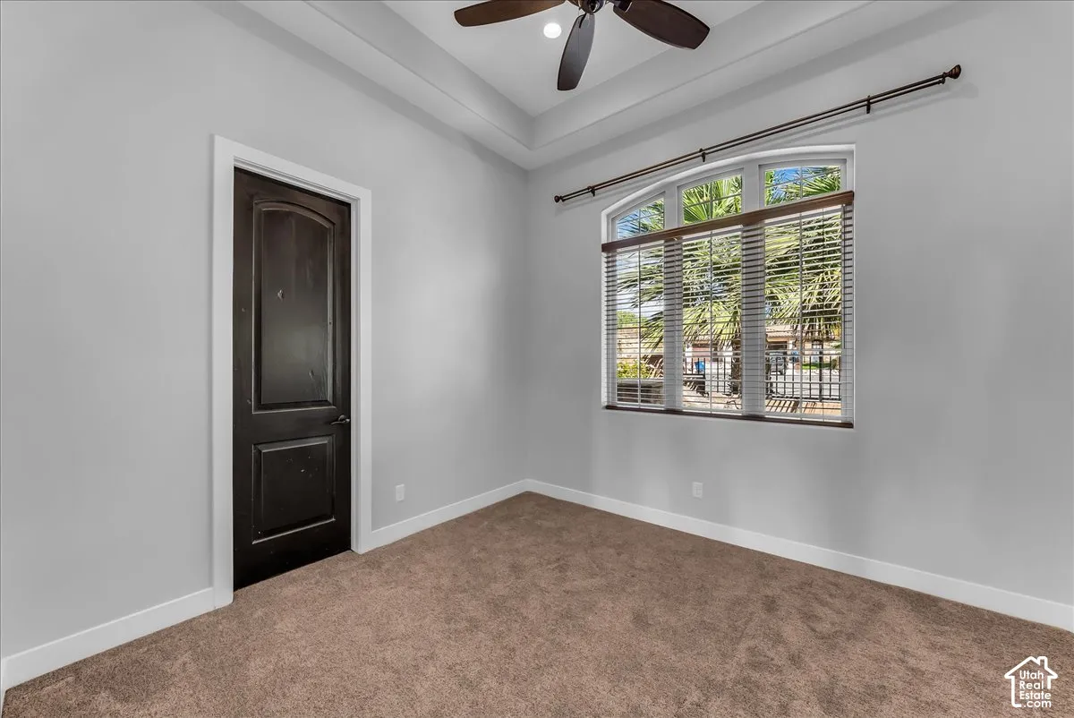 Arched window with blinds, dark wood-finish door, tray ceiling, ceiling fan, and carpeted flooring