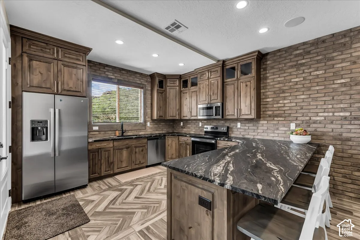 Full kitchen featuring wood cabinetry, stainless steel appliances, a stone-finish island with seating, and a brick-finish accent wall
