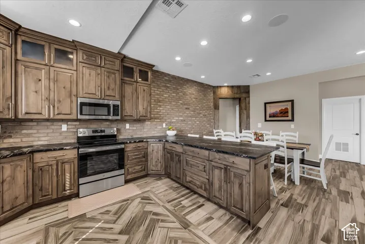Kitchen featuring wood cabinetry, stainless steel appliances, a brick-finish accent wall, dark countertops, and wood-finish flooring