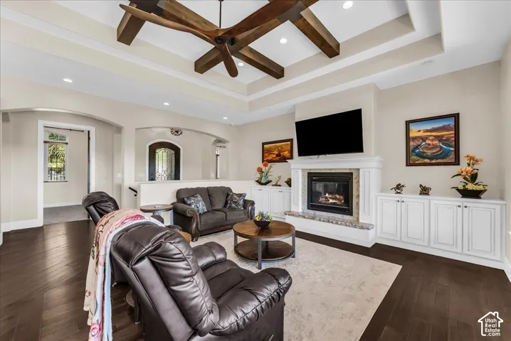 Tray ceiling with wood beam accents, recessed lighting, fireplace with stone surround and hearth, built-in cabinetry, and dark wood-finish flooring