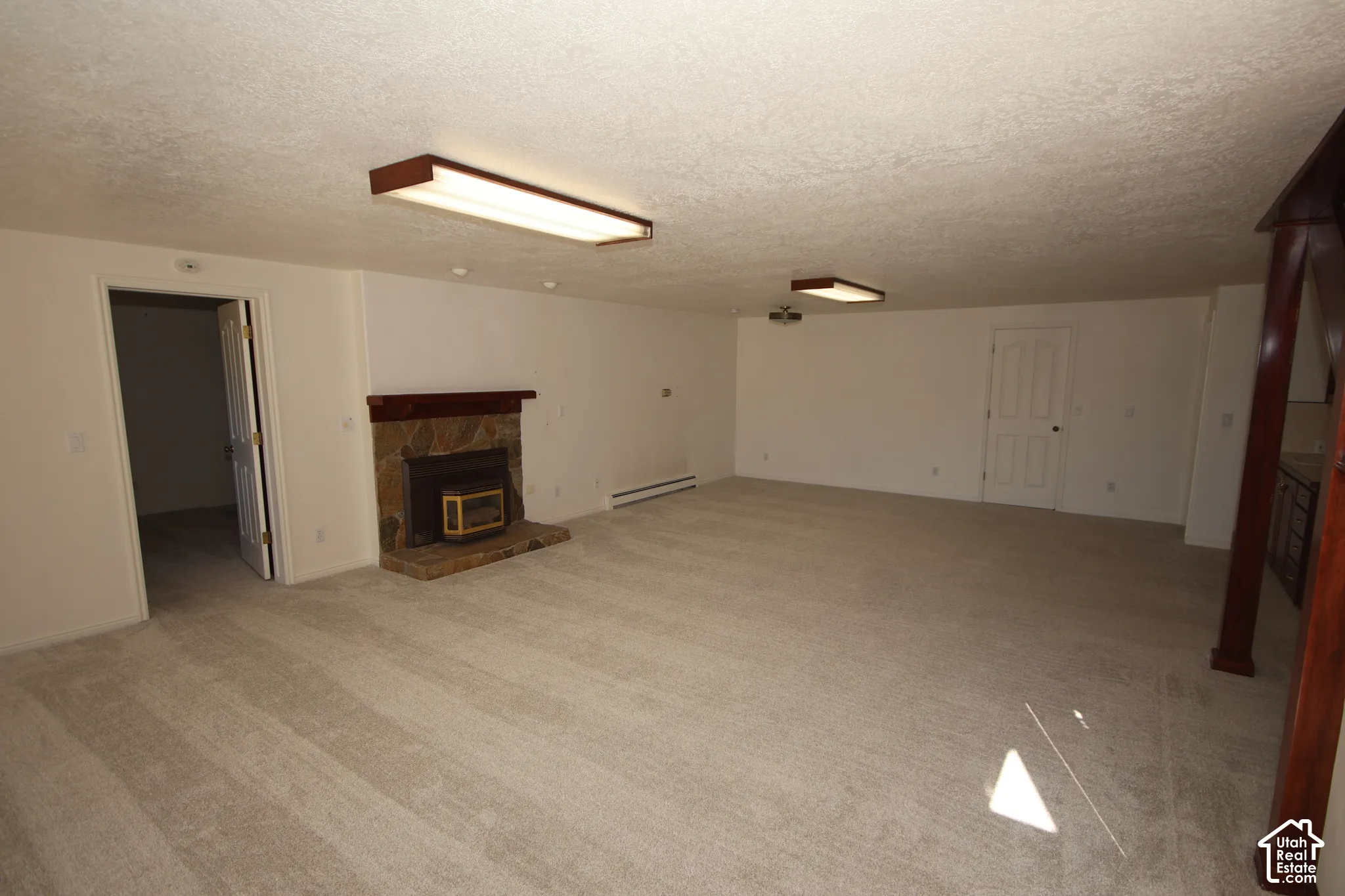 Spacious room featuring neutral carpeting, a stone-faced fireplace with a wood mantle, and bright overhead lighting