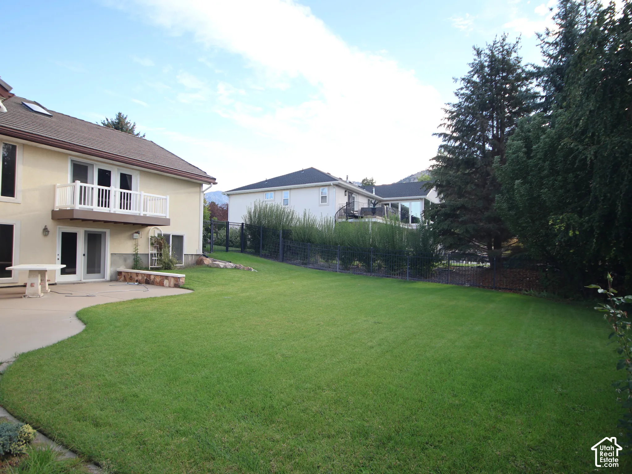 Expansive lawn area with a concrete patio, a second-story balcony with white railings, mature trees, and a low stone retaining wall
