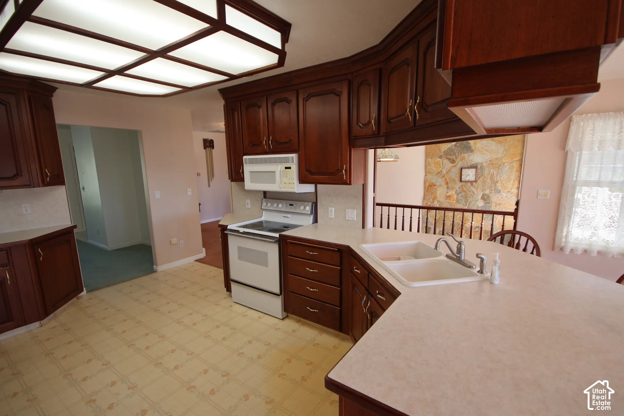 Kitchen featuring extensive wood-finish cabinetry, light-toned countertops, integrated double basin sink, white appliances, and a coffered ceiling light fixture