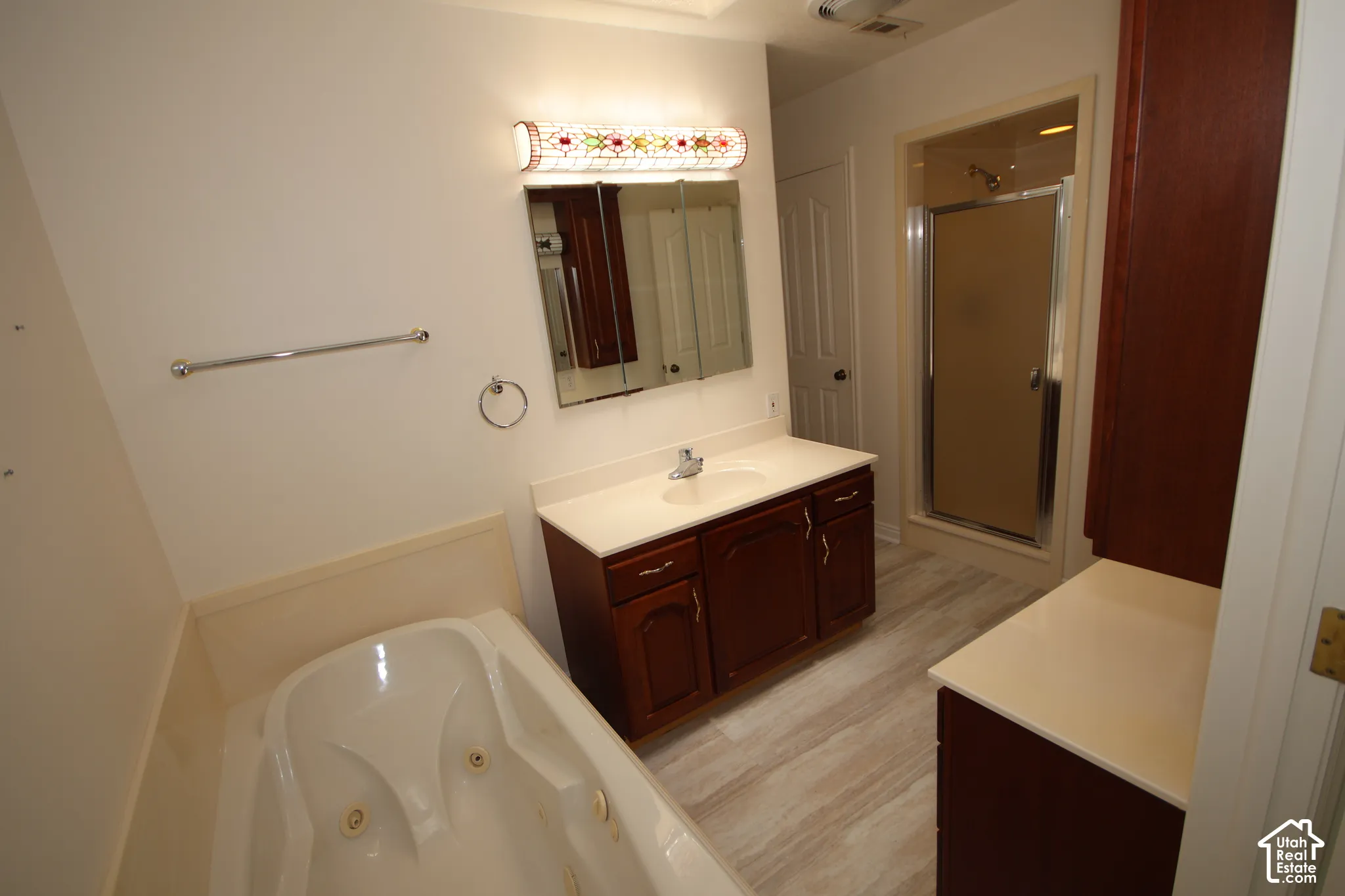 Jetted soaking tub, vanity with integrated sink, wood-finish cabinetry, walk-in shower with glass enclosure, and light-toned flooring