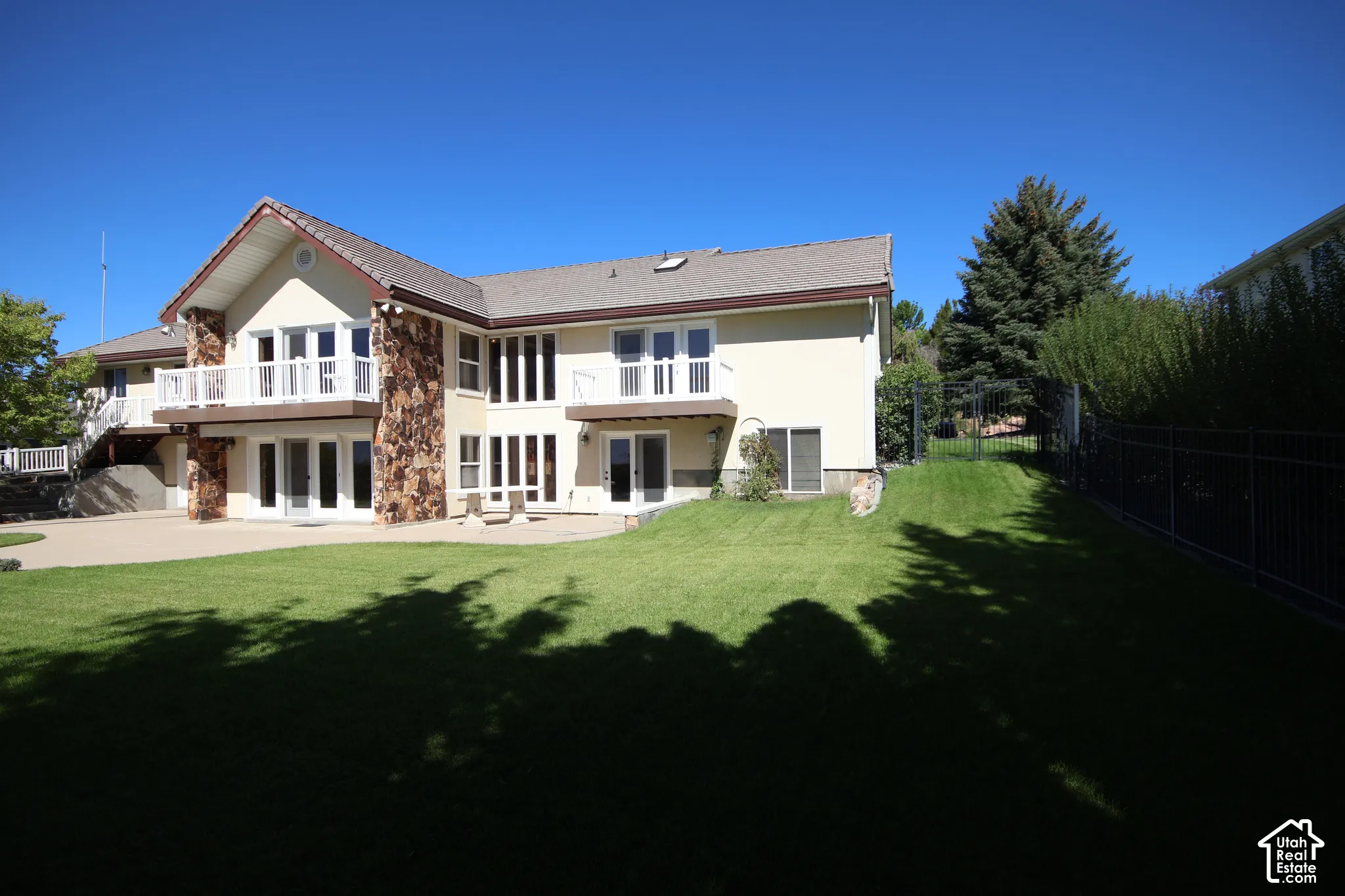 Stucco exterior with stone accents, multiple balconies with white railings, a large patio, extensive lawn, and a black wrought iron fence