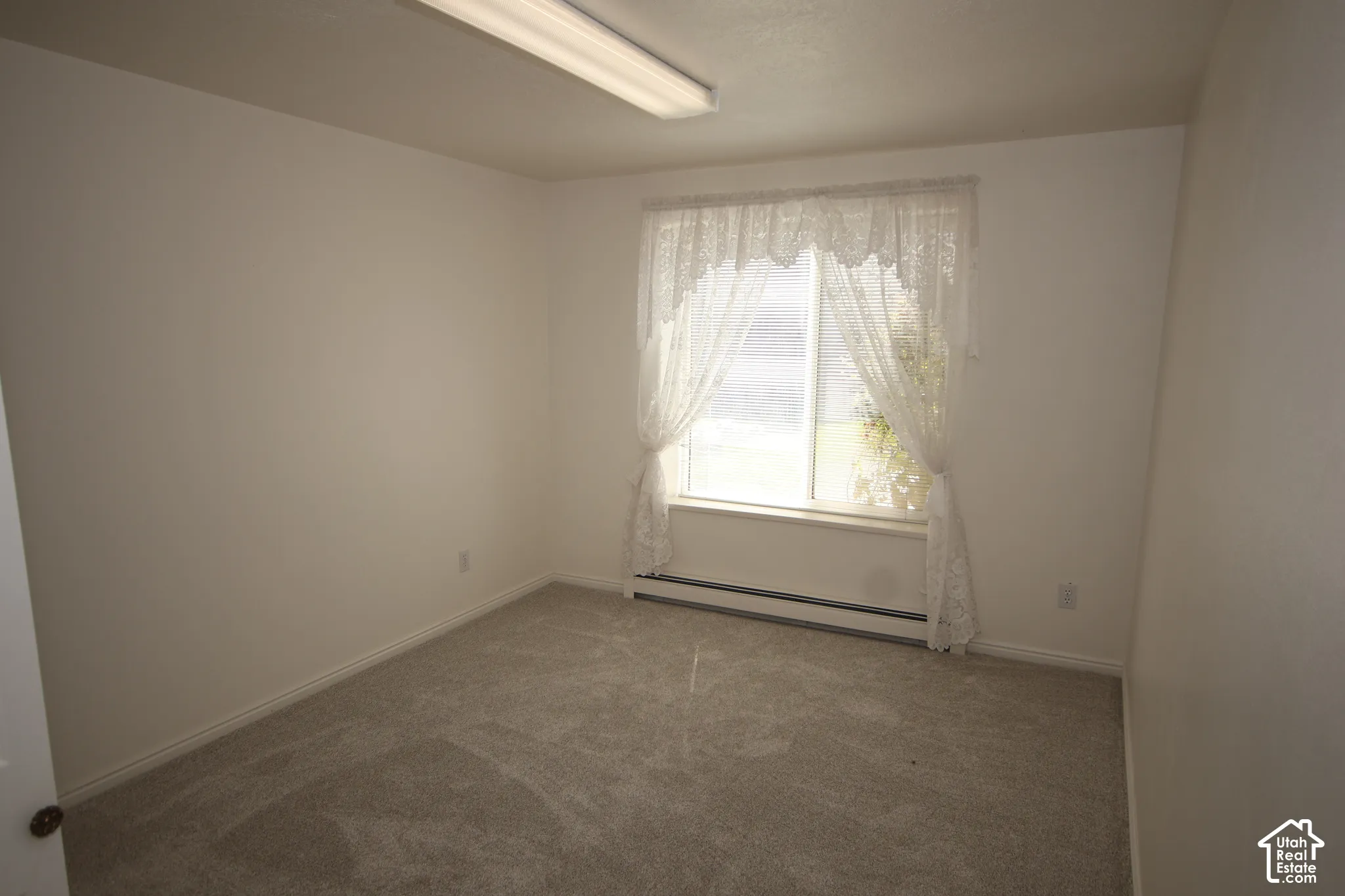 Carpeted room with a single window, white walls, and an overhead fluorescent light fixture