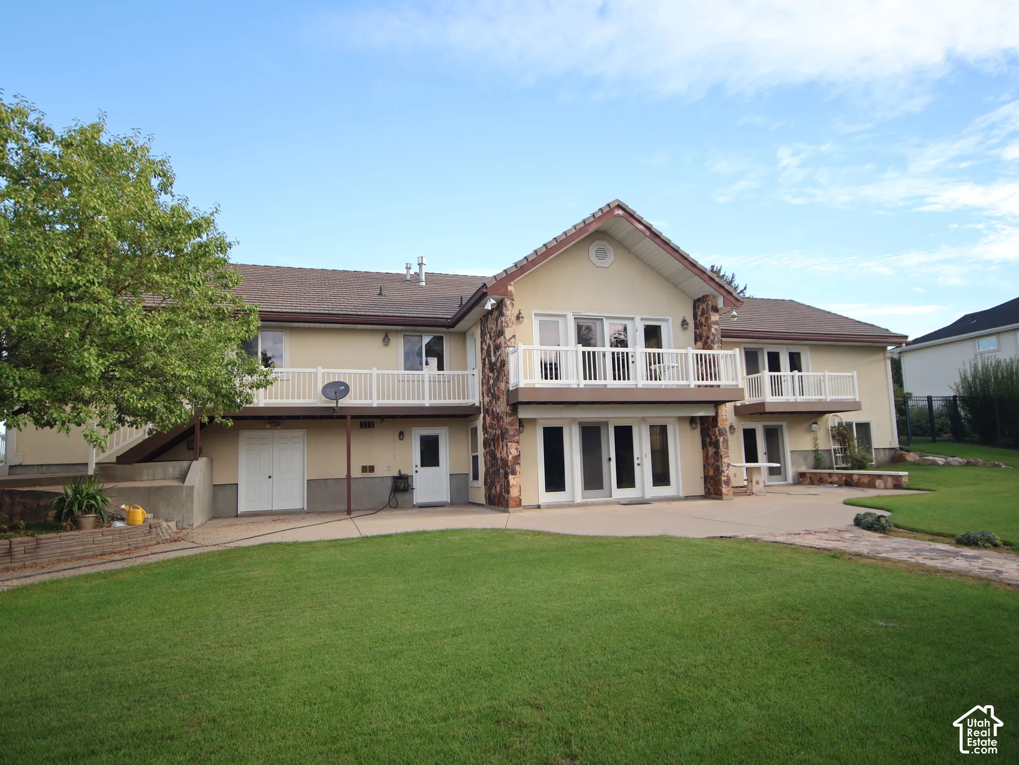 Expansive green lawn, multi-level patio, dual upper-level balconies with white railings, and a stucco exterior with stone accents