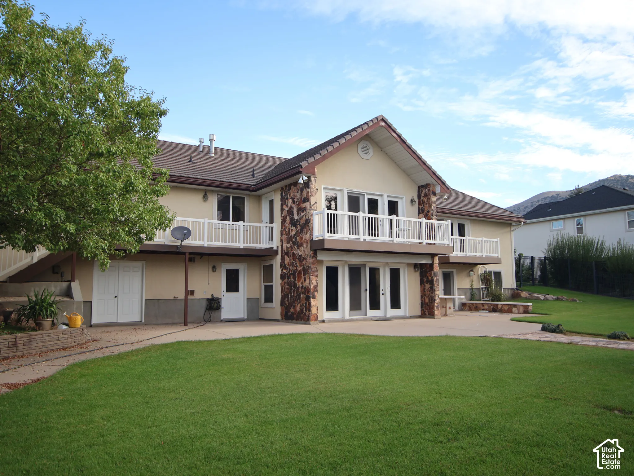 Expansive green lawn, stone patio with built-in seating, multiple walk-out doors, upper-level balconies with white railings, and stone-clad architectural accents