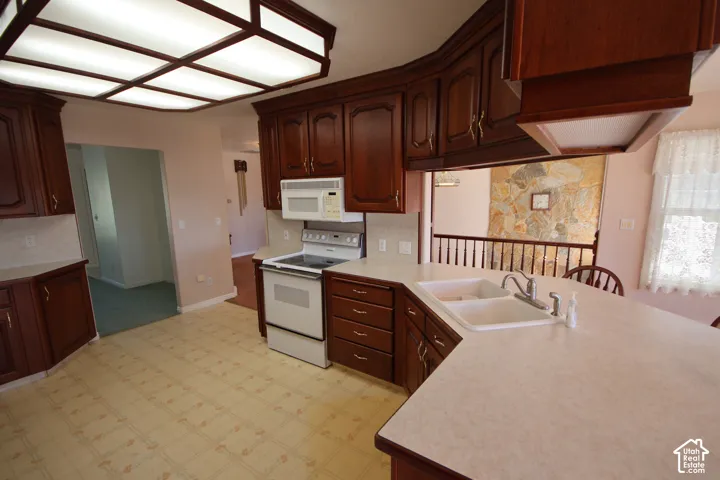 Kitchen featuring extensive wood-finish cabinetry, light-toned countertops, integrated double basin sink, white appliances, and a coffered ceiling light fixture