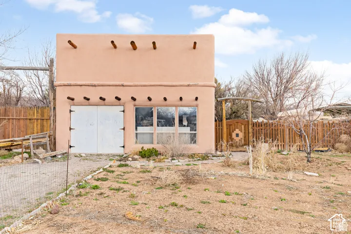 Adobe-style building featuring exposed wood vigas, a large window, double doors, and an enclosed yard with a wood fence and gate