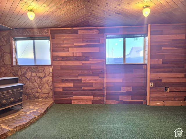 Vaulted ceiling with natural wood-plank paneling, horizontal wood-plank wall detailing, natural stone hearth, cast iron stove, and dual sliding windows