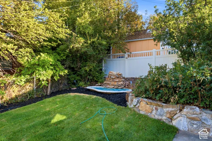 Manicured lawn with a stone-tiered water feature, surrounded by mature trees and shrubs, and a white privacy fence