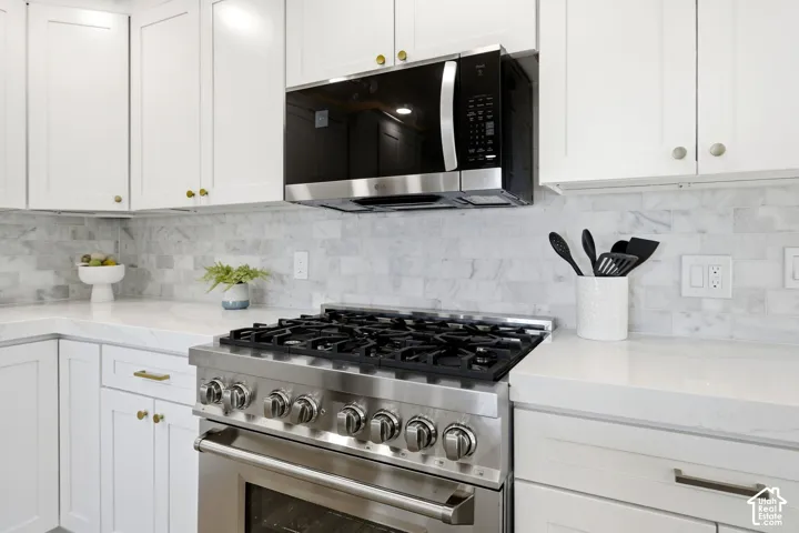 Stainless steel range with oven, custom white cabinetry with gold-tone hardware, white countertop, white tile backsplash, and built-in microwave
