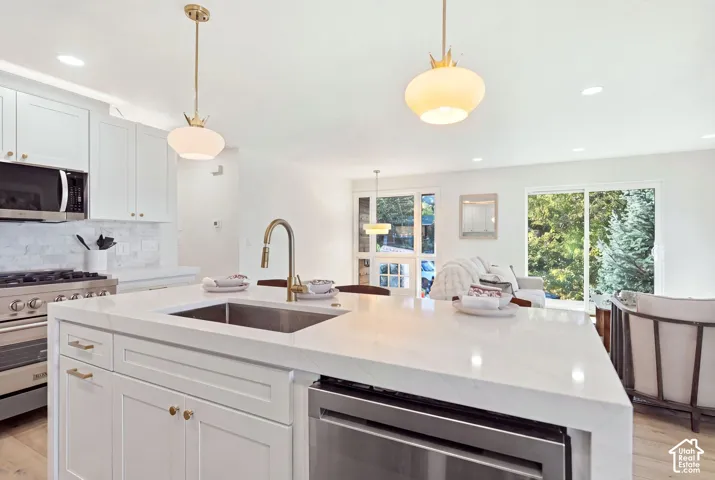 White shaker cabinetry, stainless steel appliances, brushed gold fixtures, a center island with an integrated sink, and wood-finish flooring