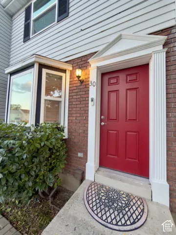 Red panel door with decorative white trim, brick facade, bay window, exterior wall sconce, and mature landscaping