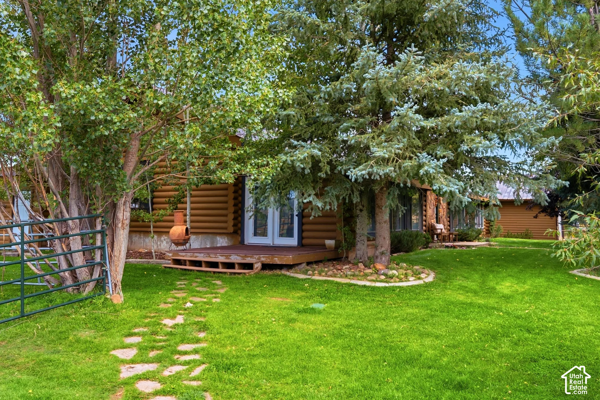 Log-style exterior with a wood-finish deck, white frame French doors, mature trees, and a stone walkway