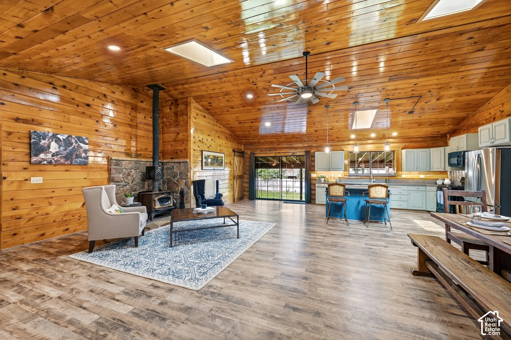 Vaulted wood-paneled ceilings, wood-finish flooring, skylights, stone fireplace with a wood-burning stove, and a kitchen with light-toned cabinetry and stainless steel appliances